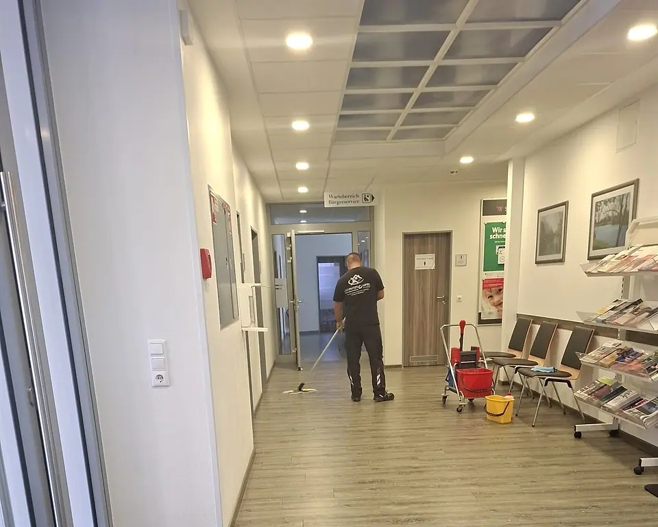 Mitarbeiter wischt den Flur einer Verwaltung, daneben steht der Reinigungswagen
